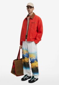 Rote Jacke mit braunem Kragen, gestreiftes Hemd, weiße Hose mit Landschaftsprint, braune Tasche mit roten Akzenten und schwarze strukturierte Sneakers.