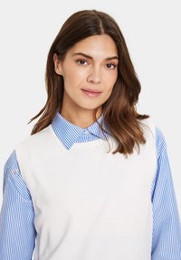 Weißer ärmelloser Pullover über einem blau-weißen gestreiften Hemd mit Kragen. Glattes Material mit klassischem Kragen und dezentem Logo-Detail am Ärmel.