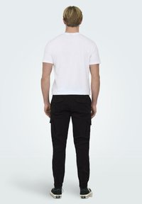 T-shirt en coton blanc associé à un pantalon cargo noir avec des poches latérales et des poignets élastiques, porté avec des baskets sombres.