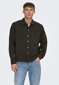 Camisa de botones marrón con un bolsillo en el pecho, botones plateados y costuras en contraste. Llevada sobre una camiseta blanca y combinada con jeans de color azul claro.