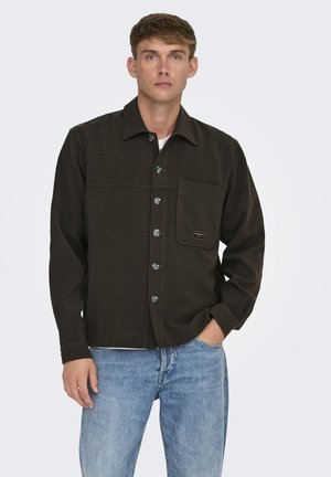 Camisa de botones marrón con un bolsillo en el pecho, botones plateados y costuras en contraste. Llevada sobre una camiseta blanca y combinada con jeans de color azul claro.
