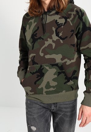 Person trägt einen grünen und braunen Camouflage-Hoodie mit Fronttasche und Kordelzug, kombiniert mit dunkelgrauen, ausgefransten Jeans.
