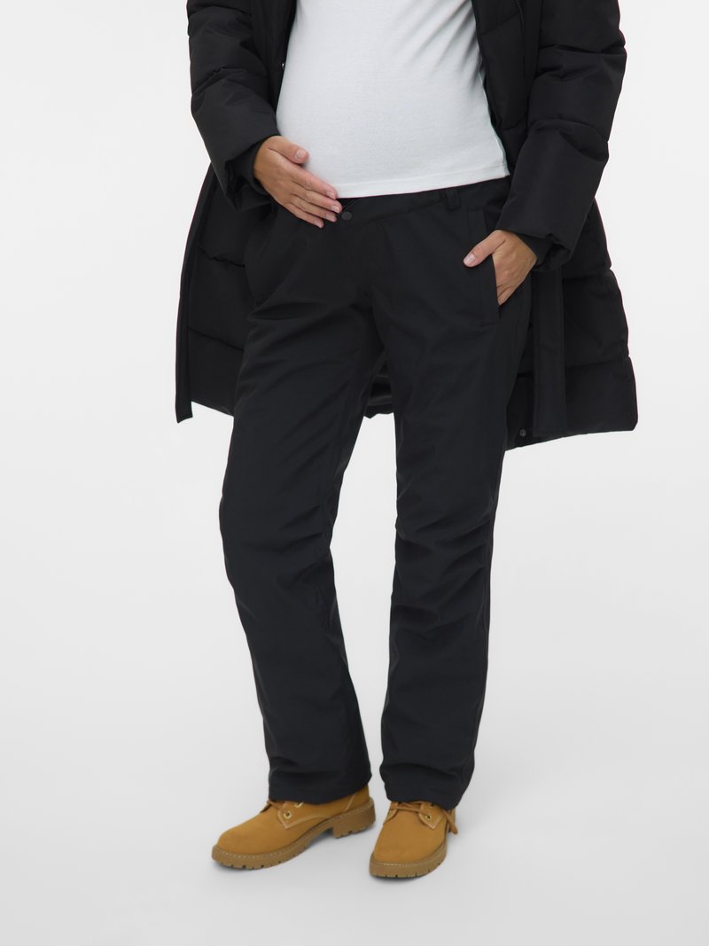 Cilvēks, kurš valkā melnus bikses, brūnas kurpes, baltu kreklu un melnu mantu, ar vienu roku uz vēdera un otru kabatā.