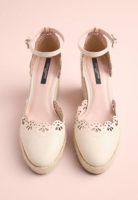 Beige espadrille shoes with scalloped cutouts, ankle strap, and woven jute sole. Smooth leather upper and soft pink lining.
