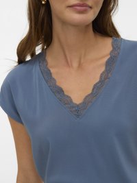 Kvinde iført en blå V-hals top med blondestrimling langs udskæringen og korte ærmer, der viser øverste del af bryst og nakke.