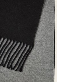 Zwarte wollen sjaal met franjes, geplaatst tegen een lichtgrijze textuurstof, die de contrasterende kleuren en materialen benadrukt.