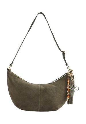 Frau mit lockigem Haar, die ein braunes, strukturiertes ärmelloses Oberteil und Hose trägt, und eine große olivgrüne Schultertasche mit Quaste und Perlen bei sich hat.