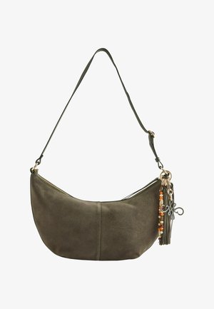 Femme aux cheveux bouclés portant un haut marron texturé sans manches et un pantalon, portant un grand sac à bandoulière vert olive avec un pompon et des perles.