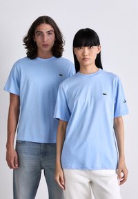 Dos personas que llevan camisetas de Lacoste de color azul claro están de pie una al lado de la otra frente a un fondo liso, mirando directamente al frente.