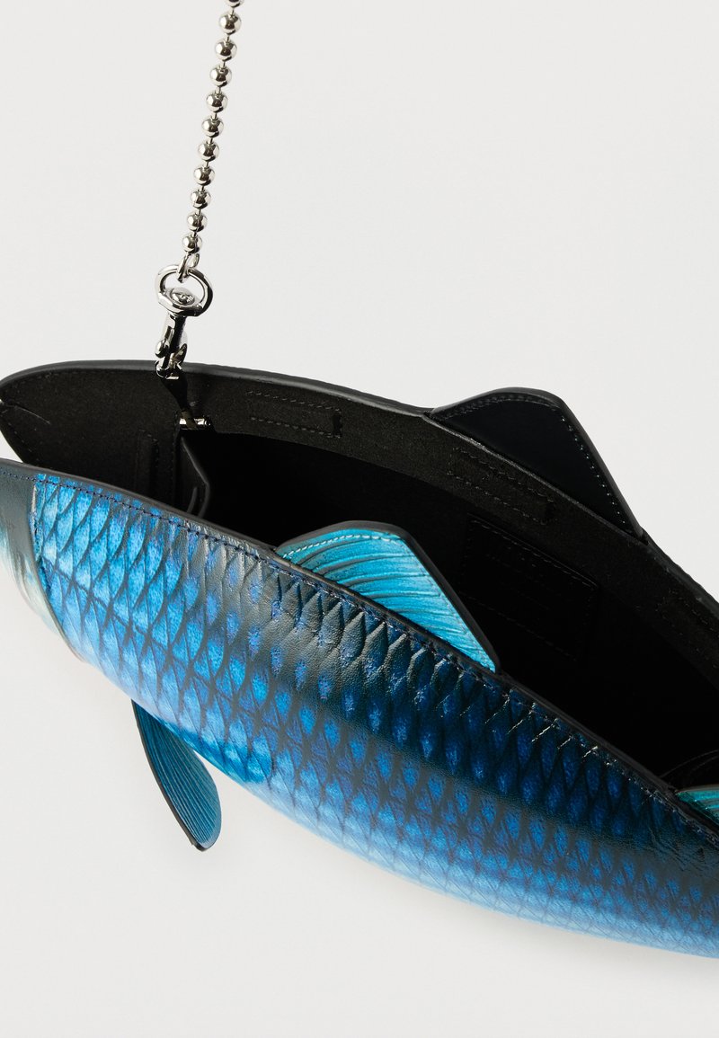 Petit sac en forme de poisson bleu avec texture d'écailles, intérieur noir, et anse chaîne argentée suspendu sur un fond blanc.