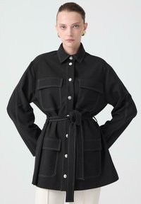 Crna oversized košulja jakna napravljena od čvrstog materijala. Sadrži srebrne dugmadi, kontrastnu bijelu šav, dvije prednje džepove i pojas koji se veže oko struka.