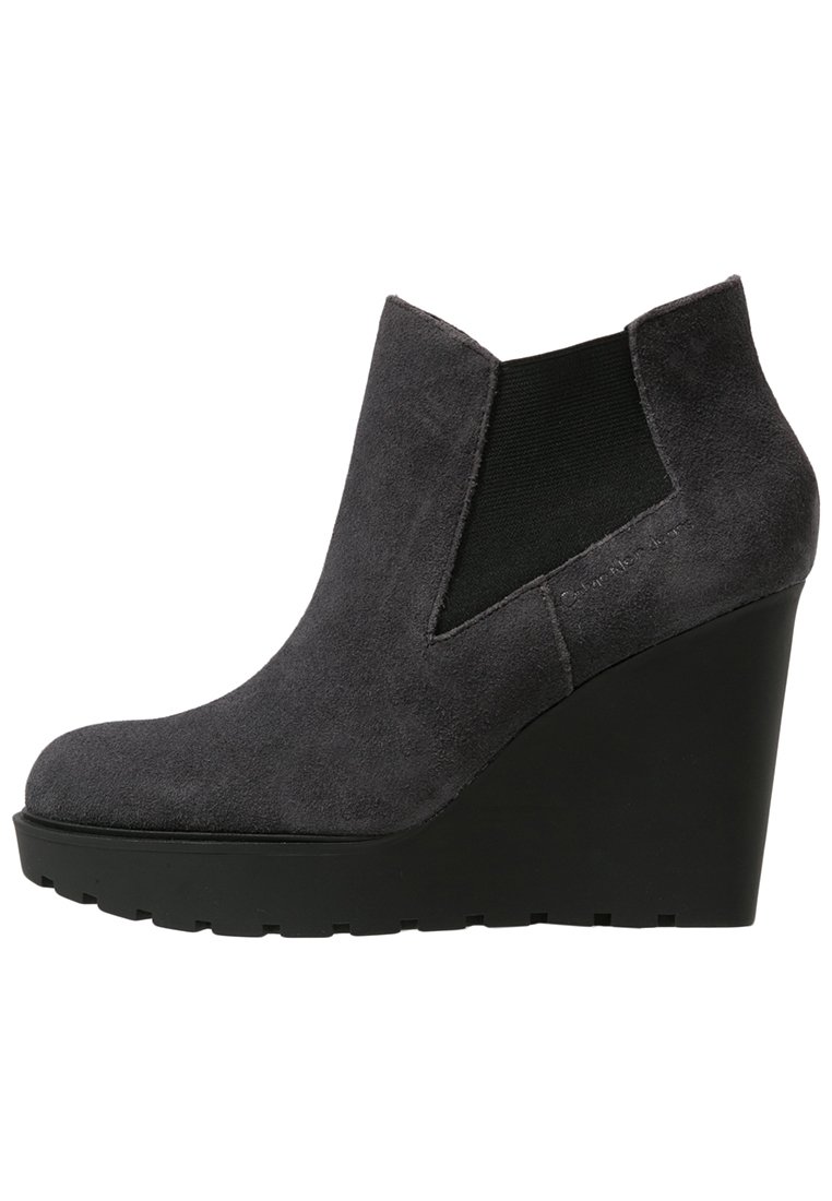 Stivale ankle grigio in suede con tacco a zeppa, punta arrotondata, pannelli elastici laterali e suola in gomma nera testurizzata. Finitura liscia con un aspetto opaco.