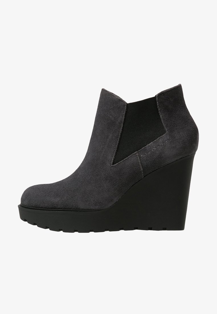 Stivale ankle grigio in suede con tacco a zeppa, punta arrotondata, pannelli elastici laterali e suola in gomma nera testurizzata. Finitura liscia con un aspetto opaco.
