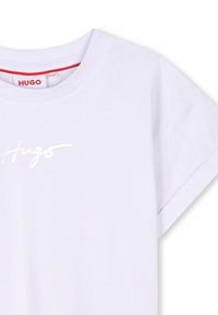 Balta T-krekla ar īsām piedurknēm, brīvs piegriezums un spīdīgs "Hugo" logo priekšpusē. Materiāls izskatās mīksts un gluds.