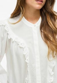 Biała bluzka z falbaniastym wzorem z przodu, z zapięciem na guziki przy szyi. Materiał wydaje się lekki i teksturowany.