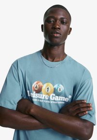 Svijetloplava pamučna majica s grafičkim printom šarenih bilijarskih kugli i natpisom "Leisure Game" u bijeloj boji. Kratki rukavi, ležeran kroj.