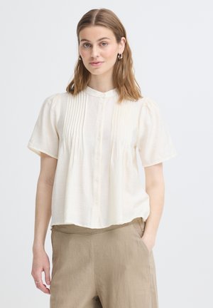 Młoda kobieta o brązowych włosach do ramion, ubrana w kremową bluzkę z krótkim rękawem z plisami i beżowe spodnie z wysokim stanem, stoi z jedną ręką w kieszeni.