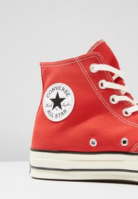 Red canvas high-top sneaker with white eyelets and laces, featuring a circular black and white "Converse All Star" patch on the ankle.