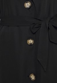 Zwarte jas met drie grote bruine schildpadknopen en een strik van stof als riem in de taille.