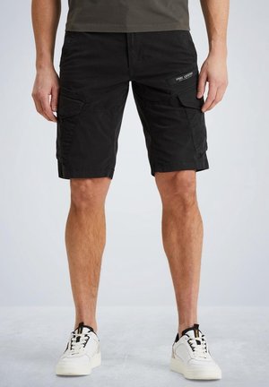 Homme portant un short cargo noir et des baskets blanches, debout devant un fond clair uni.