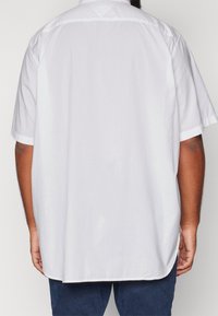Vista posteriore di una persona che indossa una camicia bianca a maniche corte e di taglio ampio, e pantaloni blu scuro, in piedi contro uno sfondo chiaro e neutro.