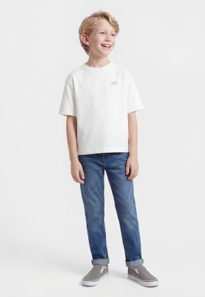 Ragazzo biondo sorridente che indossa una maglietta bianca, jeans blu arrotolati e scarpe slip-on grigie, in piedi su uno sfondo bianco.