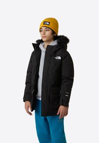 Μαύρο μπουφάν puffer με κουκούλα επενδυμένη με γούνα, που διαθέτει μπροστινό φερμουάρ, τσέπες και το λογότυπο The North Face. Φορεμένο πάνω από γκρι φούτερ με κουκούλα και μπλε παντελόνι.