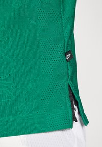 Acercamiento de tela deportiva verde transpirable con sutiles patrones en relieve y una etiqueta negra de Nike "FREAK" cerca de una abertura lateral.