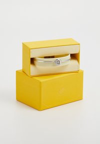 Ezüst bangle karkötő kör alakú kristály részlettel, sárga dobozban, sima textúrájú felülettel és eltávolítható selyem belsővel bemutatva.