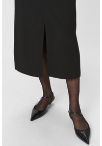 Jupe midi noire avec une fente à l'avant, associée à des chaussures noires à bout pointu avec des fines brides à la cheville. Des collants noirs transparents sont portés.