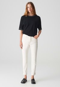 Schwarzes kurzärmeliges Shirt mit weißem Saum, kombiniert mit enganliegenden weißen Jeans mit seitlichen Schlitzen und schwarzen Slippern auf einer glatten Oberfläche.