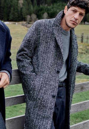 Homme portant un pardessus gris à motifs et un pull gris, appuyé contre une clôture en bois avec un champ herbeux et des arbres en arrière-plan.