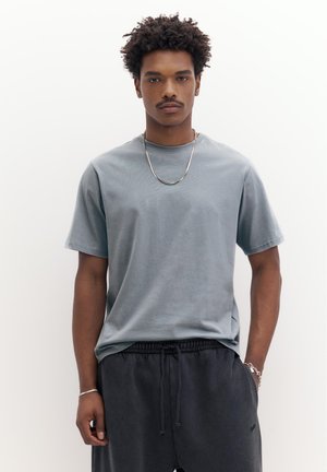 Junger Mann mit lockigem Haar, der ein hellgraues T-Shirt, dunkle Hosen mit Kordelzug und silbernen Schmuck trägt, vor einem schlichten weißen Hintergrund steht.