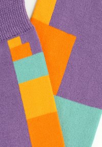 Kötött zoknik, amelyek lila alapszínnel rendelkeznek, narancssárga és türkiz színű geometrikus mintákkal, kiemelkedő színblokkokkal és struktúrált szövésű kivitelezéssel.