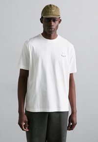 Λευκό μπλουζάκι από βαμβάκι με κοντά μανίκια και στρογγυλή λαιμόκοψη, που διαθέτει κεντητό σκοτεινό μπλε χαμογελαστό πρόσωπο στο στήθος.