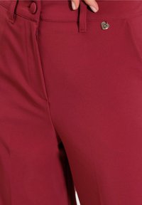 Burgunderfärgade byxor i slät tyg, med en knapp och en liten hjärtformad detalj i metall vid midjan.