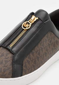 Μαύρα και καφεδιά sneakers με επάνω από ψεύτικο δέρμα, ακανόνιστο σχέδιο, χρυσό φερμουάρ και λευκή καουτσούκ σόλα. Περιλαμβάνει διακοσμητικό λογότυπο στο φερμουάρ.