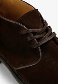 Zapato de ante marrón con punta redondeada, que presenta cordones oscuros texturizados y un forro de cuero suave, colocado sobre un fondo neutro.