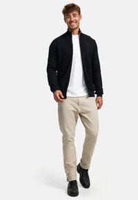 Chaqueta negra con cremallera y patrón de chevron texturizado, llevada sobre una camiseta blanca, combinada con pantalones beiges y botas negras.
