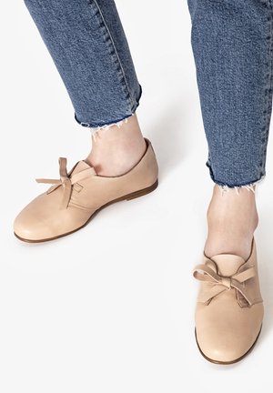 Beige lædersko med rund tå og sløjfedetalje foran, parret med blå jeans med frynset kant. Skoene har en lav brun sål.