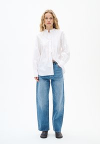 Weißes Hemd mit langen Ärmeln und gefaltetem Frontdesign, kombiniert mit hellblauen weit geschnittenen Jeans und dunklen Ankle Boots. Sauberer, minimalistischer Look.
