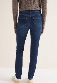 Ciemnoniebieskie skinny jeans z wysokim stanem, z dwoma tylnymi kieszeniami i gładką teksturą denimu. Noszone z teksturowanym brązowym swetrem.
