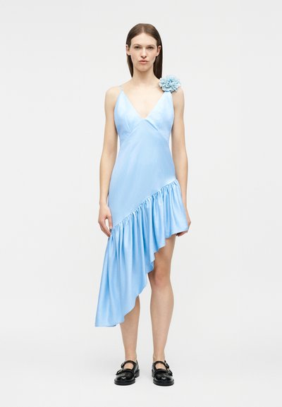 Γυναίκα που φοράει ανοιχτό μπλε ασύμμετρο φόρεμα με βολάν στο τελείωμα και floral λεπτομέρεια στον ώμο, συνδυασμένο με μαύρα παπούτσια Mary Jane.