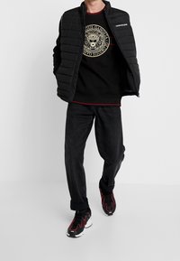 Chaleco acolchado negro con un cuello alto sobre un sudadera negra con un gráfico de león. Jeans oscuros y zapatillas negras con acentos rojos.