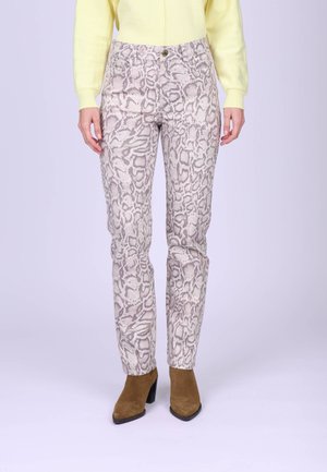 Personne portant un pantalon à imprimé serpent beige et marron, un pull jaune et des bottines en suède marron à talons, debout devant un fond uni.