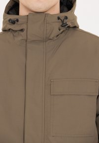 Brun huvjacka i texturerad tyg, med dragkedja fram, justerbar snörning i kragen och en ficka på bröstet.