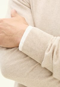 Beige Strickpullover mit gerippten Bündchen. Der Stoff fühlt sich weich an und hat eine glatte Textur. Das Handgelenk ist sichtbar und zeigt die Handdetails im Kontrast zum Ärmel.