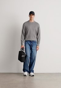 Maglietta grigia con maniche lunghe abbinata a pantaloni blu larghi, sneakers bianche e una borsa nera. Lo sfondo è semplice e di colore chiaro.