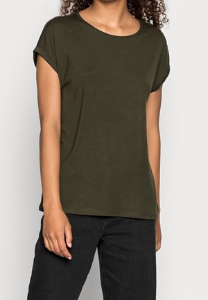 Femme portant un t-shirt à manches courtes vert foncé avec revers roulés et un pantalon noir, vue du cou jusqu'aux hanches sur un fond uni.