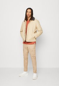 Chaqueta acolchada beige con un cuello oscuro, camisa color óxido debajo y pantalones cargo beige. Zapatillas blancas completan el atuendo.